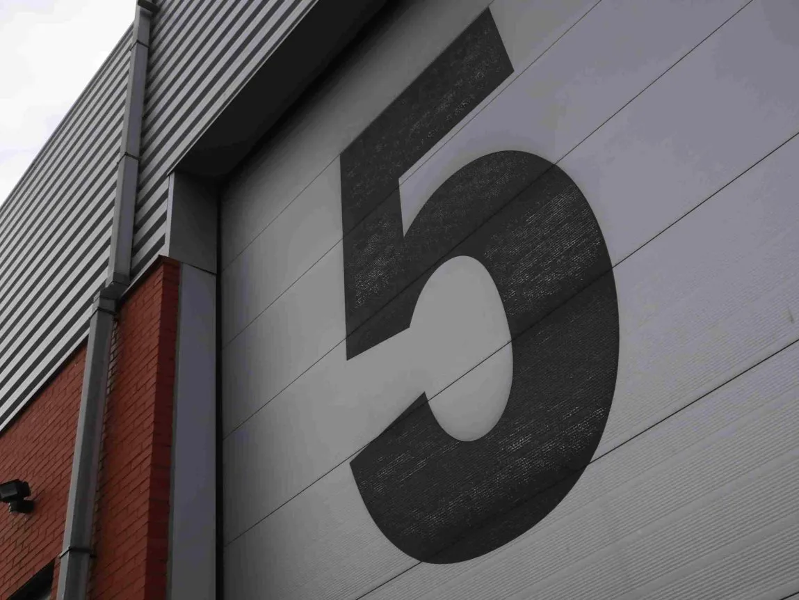 Een groot zwart cijfer 5 is geschilderd op een grijze industriële garagedeur; aan de linkerkant zijn een deel van een rood bakstenen gebouw en een metalen gevelbekleding te zien.