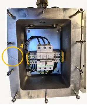 Een metalen elektriciteitsaansluitdoos met draden die zijn aangesloten op een klemmenblok binnenin. De doos is aan de randen voorzien van bouten en heeft aan de linkerkant een gele cirkel met het cijfer 4 erop.
