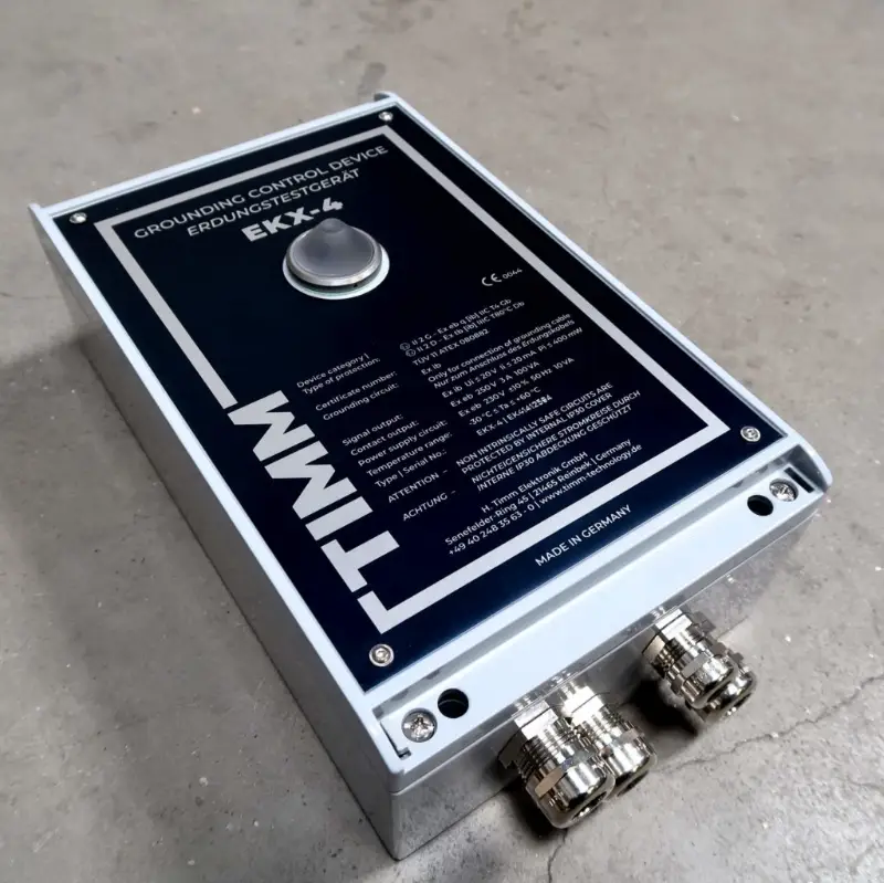 Een rechthoekig TIMM-aardingsregelapparaat (EKX-4) met een metalen knop en aansluitingen, geplaatst op een grijs betonnen oppervlak. Op het zwarte etiket staat technische tekst in meerdere talen.
