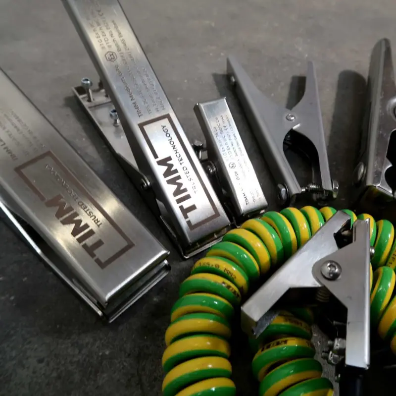 Op een grijs oppervlak liggen verschillende metalen aardingsklemmen en groen-gele, opgerolde aardingskabels. Op de klemmen staat ‘TITAM’ en nog wat andere tekst.