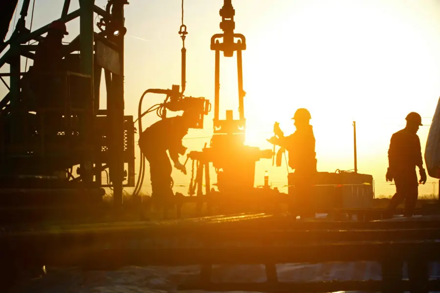 Silhouetten van werknemers die bij zonsondergang machines bedienen op een booreiland, waarbij het felle zonlicht scherpe schaduwen werpt en de contouren van de apparatuur en de figuren doet uitkomen.