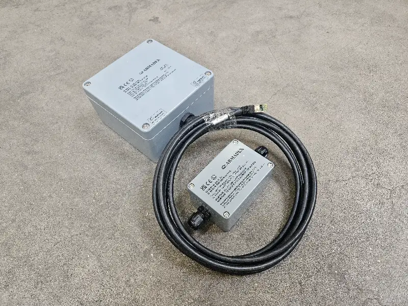 Twee grijze elektronische kastjes op een betonnen ondergrond; één groot, één klein. Een dikke zwarte kabel verbindt de twee, met aan één uiteinde een stekker met een groene punt. Op beide kastjes zitten etiketten met technische gegevens in het Japans.