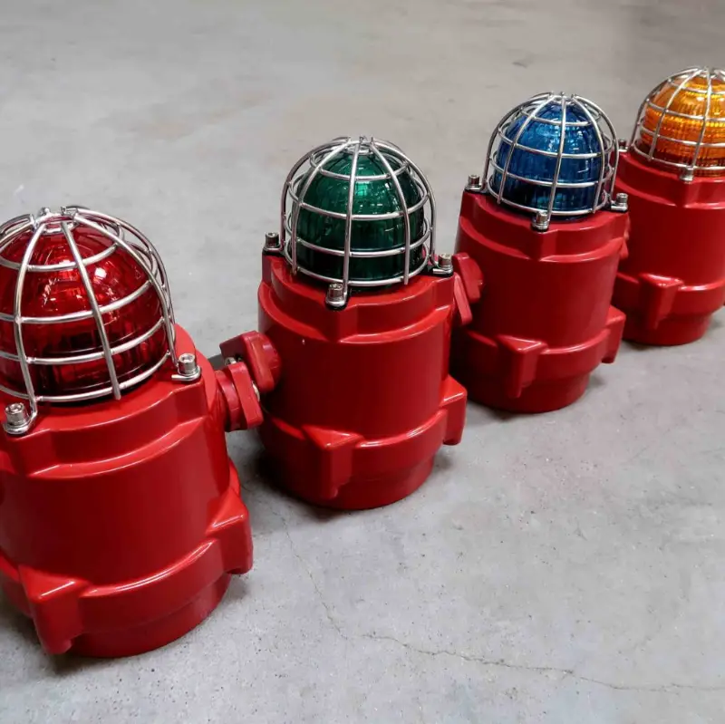 Vier rode industriële waarschuwingslampen met metalen omhulsels, elk met een lens in een andere kleur: rood, groen, blauw en geel, staan op een rij op een grijs betonnen oppervlak.