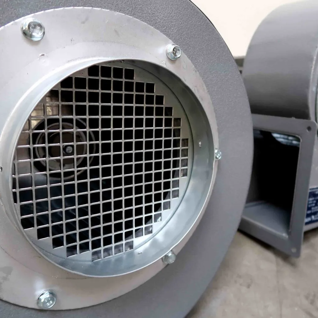Close-up van een metalen industriële ventilator of blazer met een beschermrooster over de ronde luchtinlaat, geplaatst op een gladde, lichtgekleurde vloer.