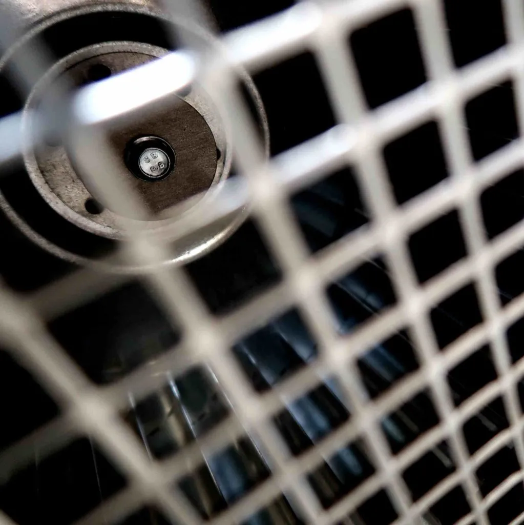 Close-up van een metalen rooster op de voorgrond, met op de wazige achtergrond een rond voorwerp waarin een klein lampje of een sensor te zien is, wat doet denken aan een industriële of mechanische omgeving.