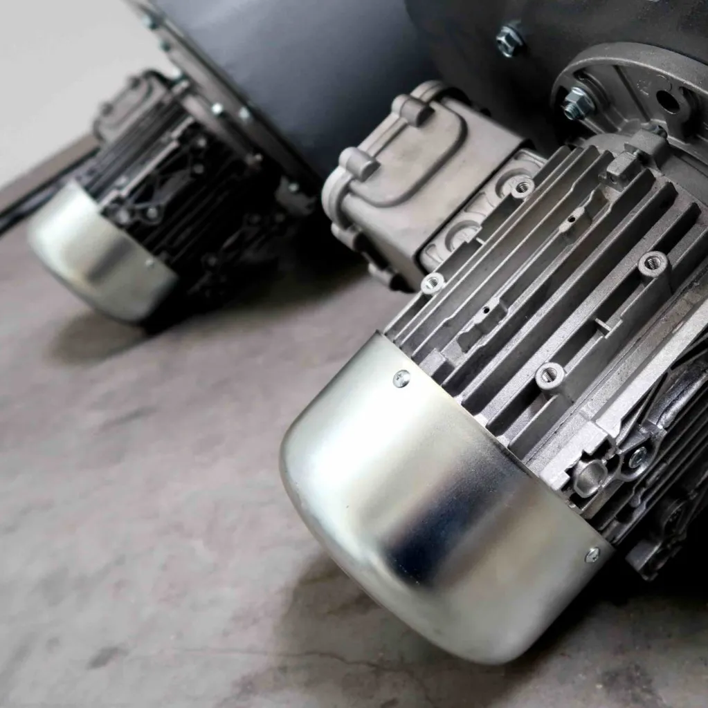 Close-up van twee industriële elektromotoren met metalen behuizingen, gemonteerd op een betonnen vloer, waarop de gedetailleerde koelribben en de beschermkappen aan de uiteinden te zien zijn.