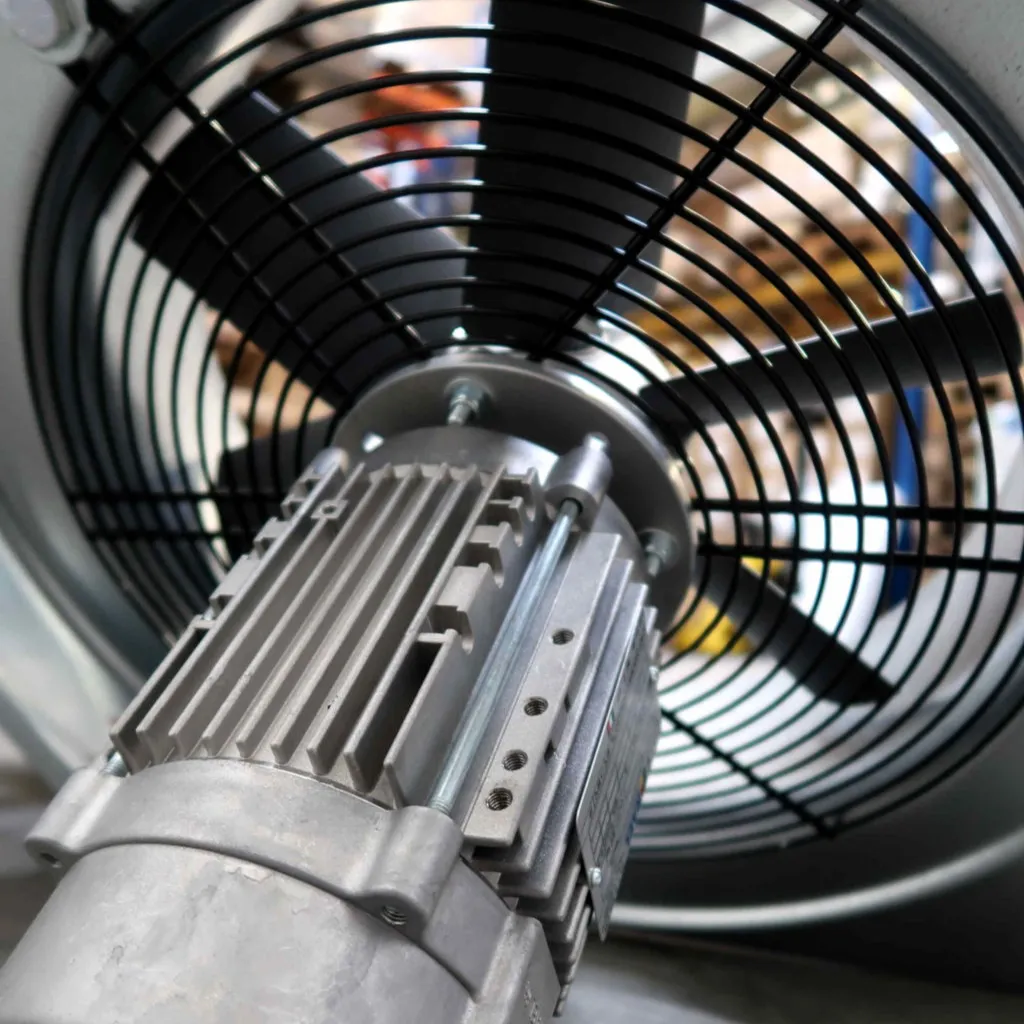 Close-up van een industriële elektromotor die is bevestigd aan een grote metalen ventilator met zwarte bladen en een beschermrooster, waarschijnlijk bedoeld voor ventilatie of koeling.