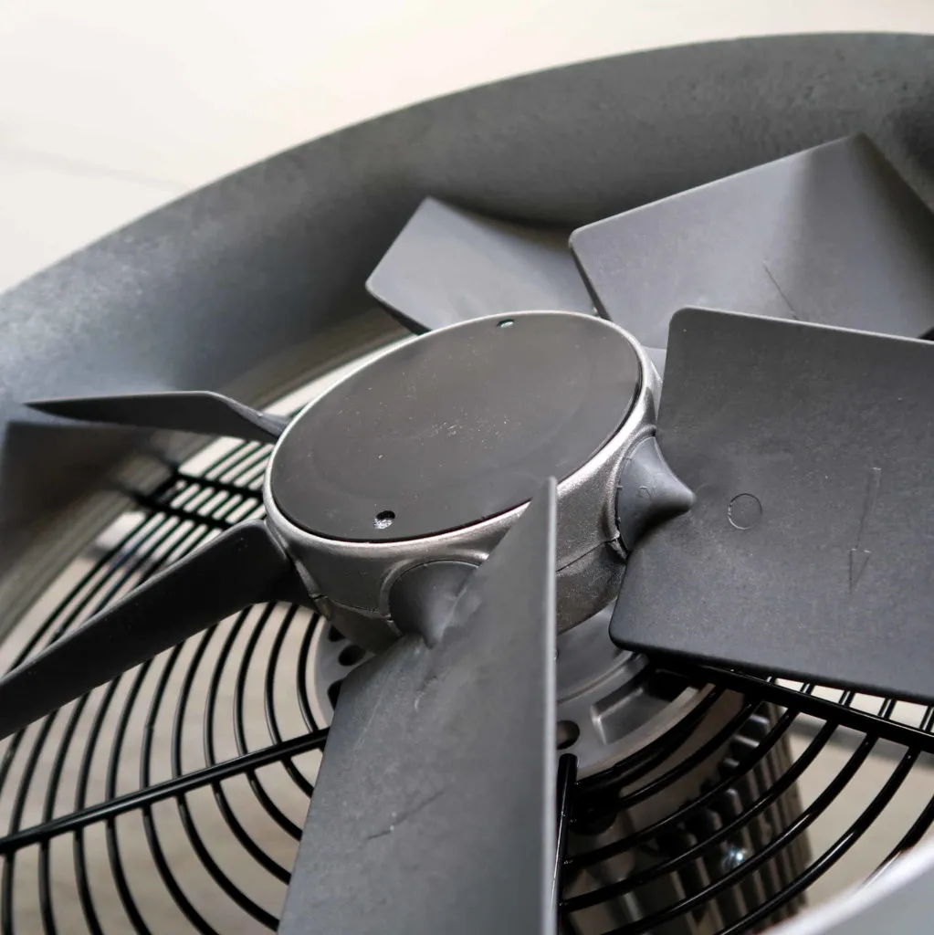 Close-up van een moderne zwarte metalen ventilator met vijf bladen, waarbij de centrale naaf en een deel van het beschermrooster op de achtergrond te zien zijn. De afbeelding benadrukt het industriële ontwerp en de textuur van de ventilator.