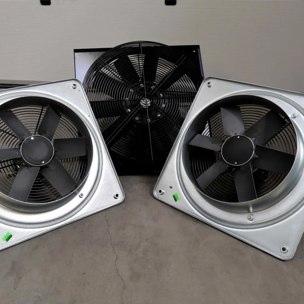 Op een betonnen vloer staan drie industriële ventilatoren met metalen frames en zwarte bladen; twee staan aan de voorkant schuin naar elkaar toe gericht, terwijl er één rechtop achter hen staat.