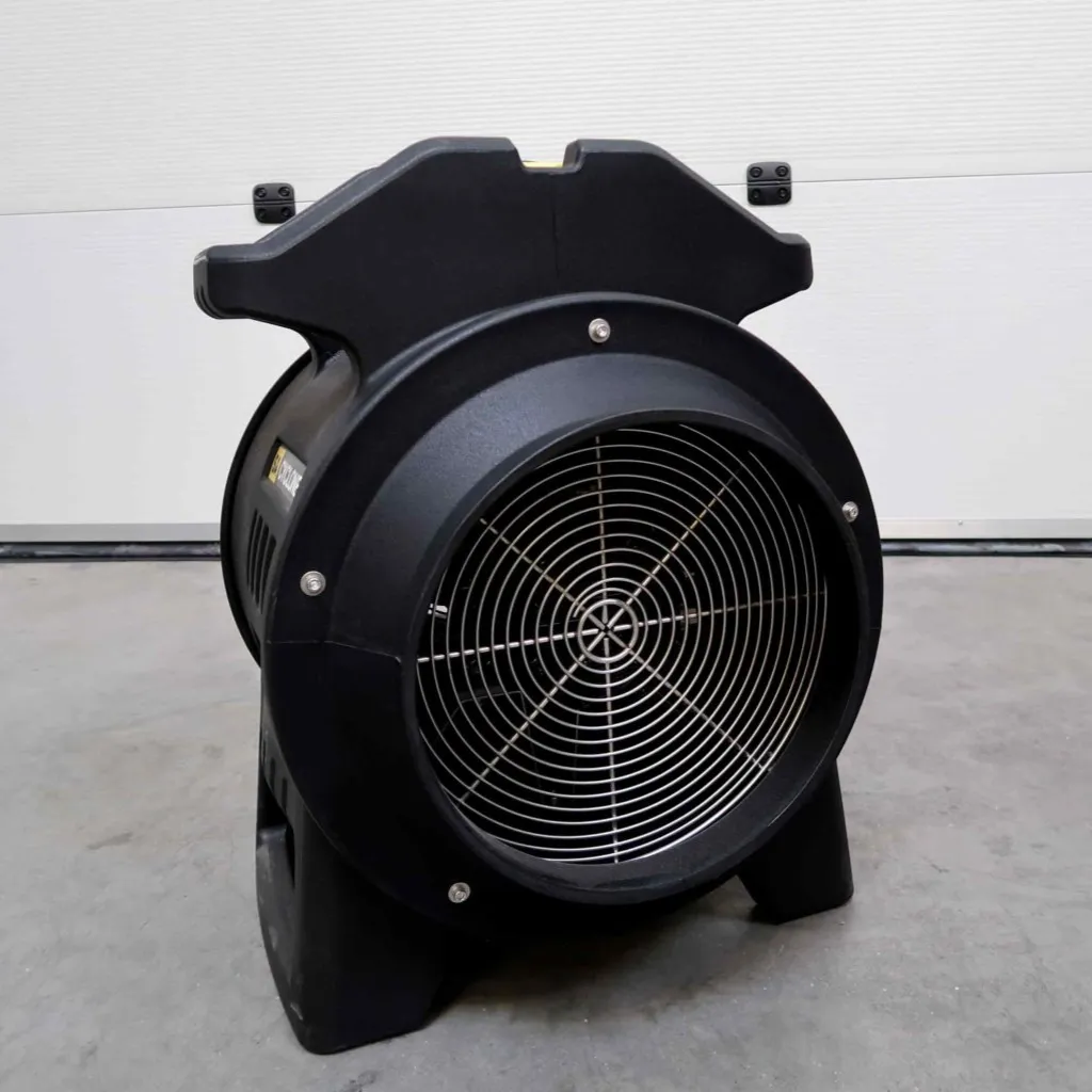 Op een grijze betonnen vloer, tegen een witte muur, staat een grote zwarte industriële ventilator met een metalen rooster.