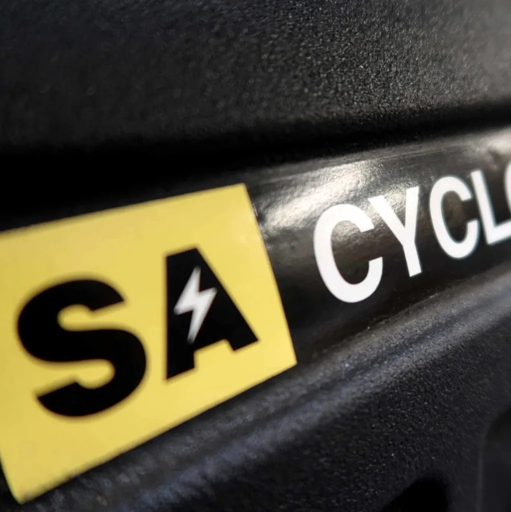 Close-up van een etiket op een zwart oppervlak. Op het etiket staat een geel vierkant met de letters SA en een bliksemschicht, gevolgd door een gedeeltelijk witte tekst: CYCLO.