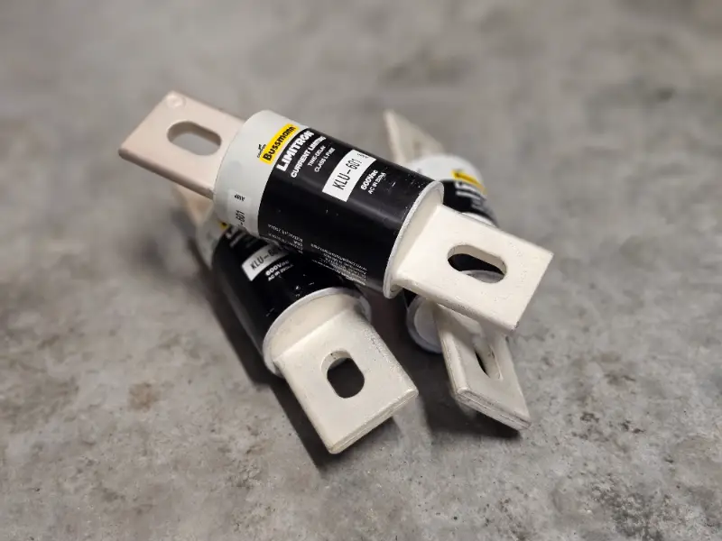 Er liggen drie cilindervormige elektrische zekeringen met zwarte etiketten en grote metalen aansluitingen op een grijs, gestructureerd oppervlak. De aansluitingen hebben rechthoekige gaten en de zekeringen liggen in een losse stapel.
