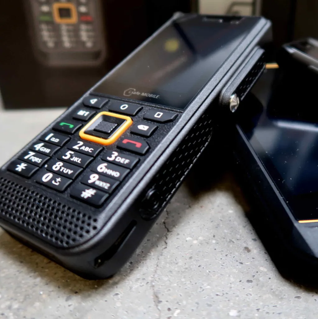 Close-up van een robuuste zwarte mobiele telefoon met fysieke knoppen en een gestructureerd oppervlak, die op een grijs oppervlak ligt naast een ander soortgelijk toestel.