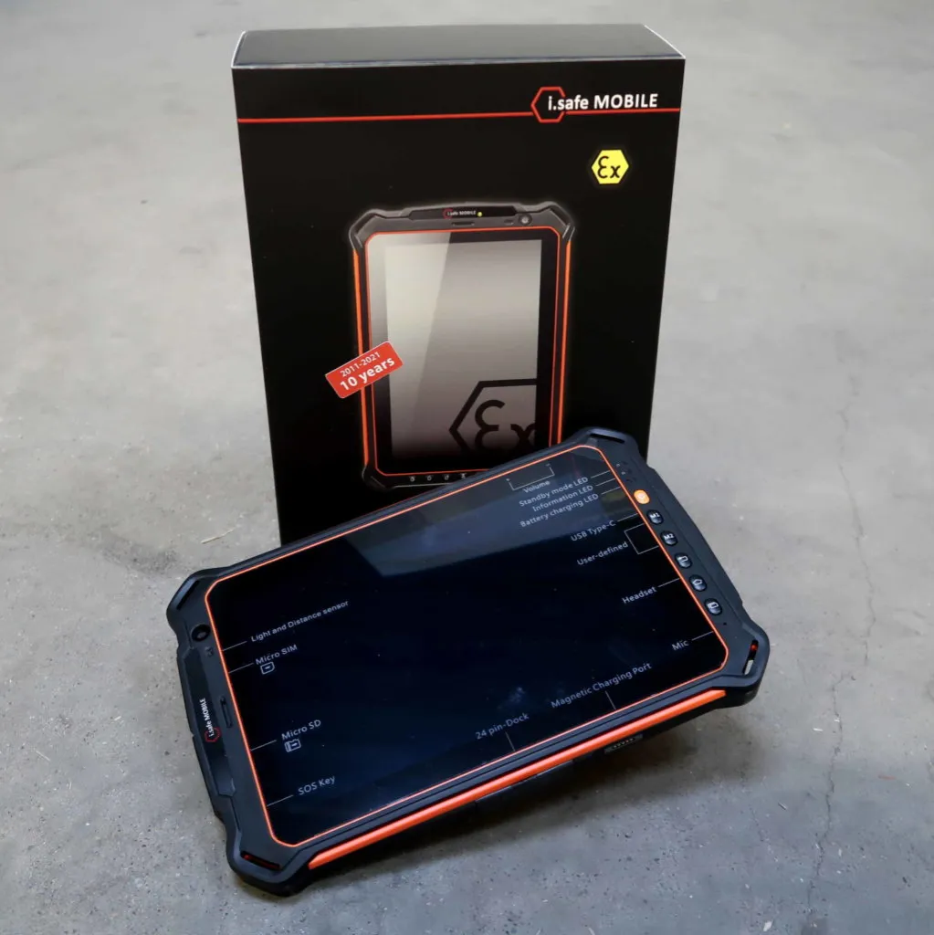 Een robuuste tablet met een behuizing met oranje rand staat tentoongesteld voor de zwarte verpakkingsdoos met het opschrift i.safe MOBILE. Het apparaat staat op een grijs oppervlak.