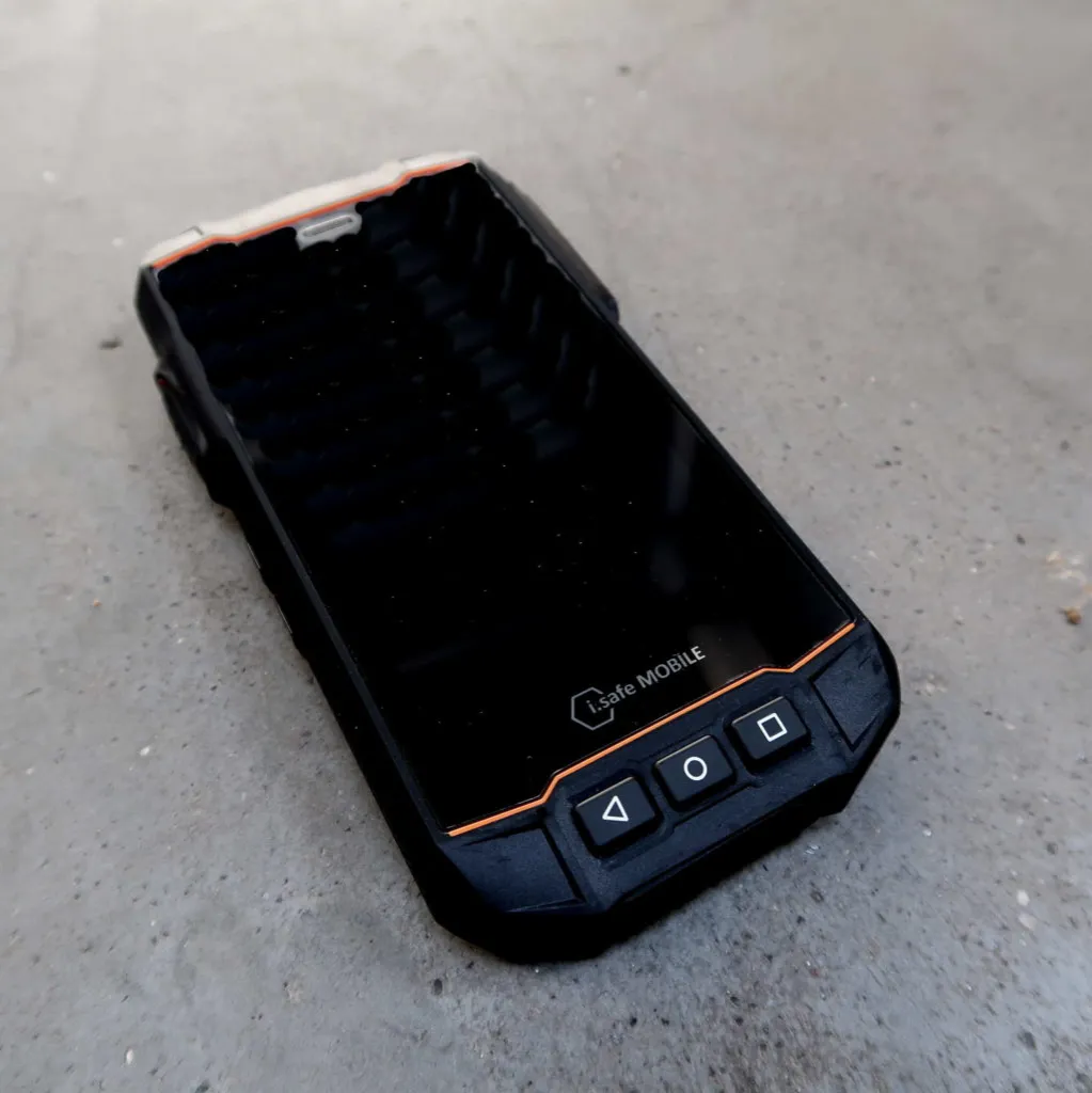 Op een grijs betonnen oppervlak ligt een robuuste zwarte smartphone met oranje accenten en drie fysieke knoppen. Op het scherm van de telefoon is het Logic MOBILE-logo te zien.