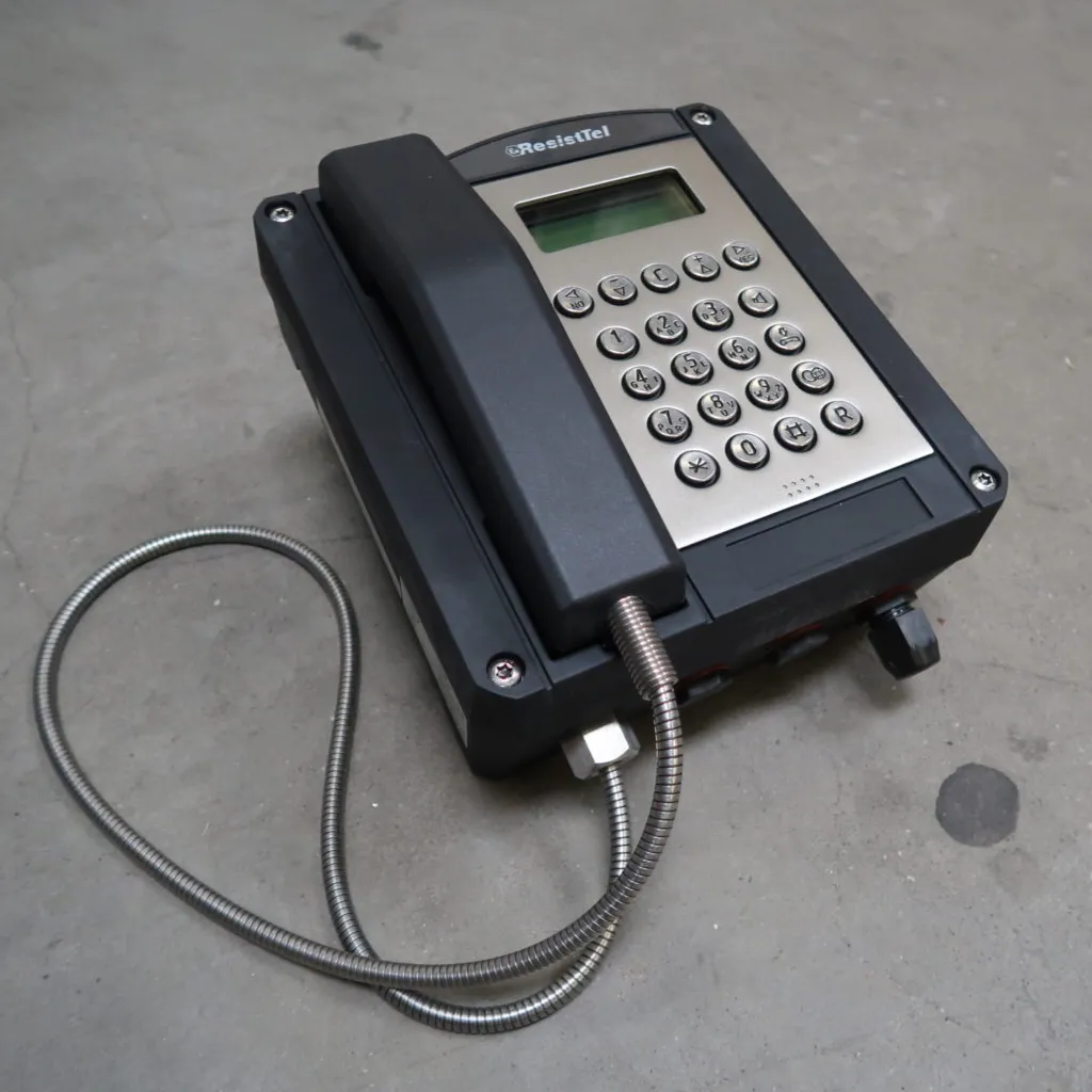 Op een grijze betonnen vloer staat een robuuste, zwart-zilveren industriële vaste telefoon met een metalen spiraalkabel.