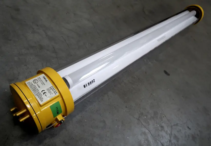 Op een grijze betonnen vloer ligt een industriële tl-armatuur met een gele kap en een doorzichtige beschermkap. Aan één uiteinde van de armatuur bevinden zich labels en aansluitingen.