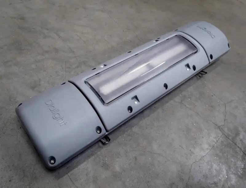 Op een betonnen vloer staat een grijze industriële LED-lamp met een doorzichtige kap. Op de behuizing is het woord ‘DrilLight’ te lezen.