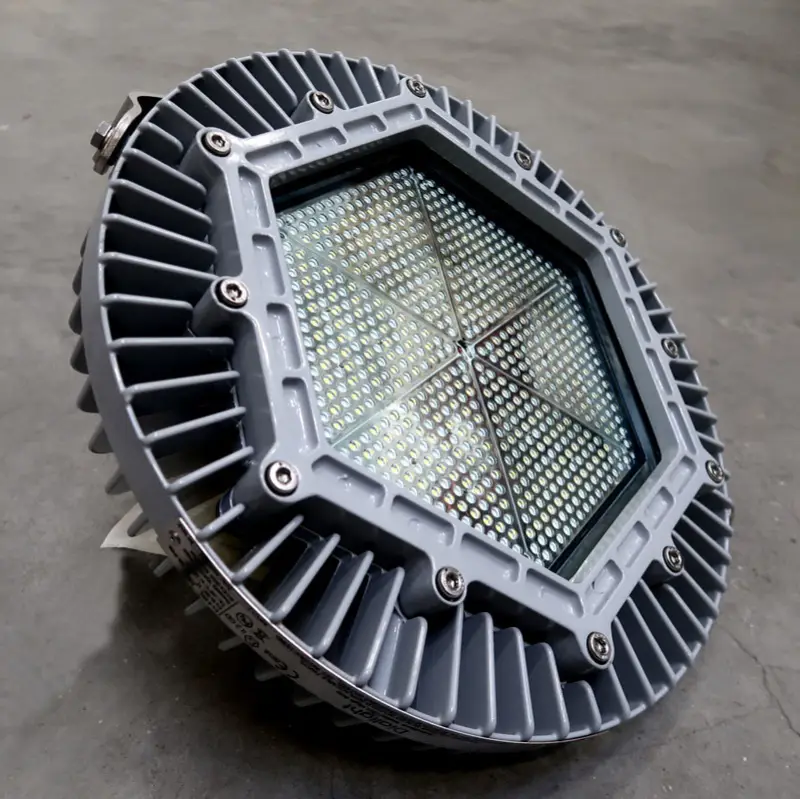 Een ronde, grijze industriële LED-lamp met een zeshoekige glazen voorkant en koelribben, geplaatst op een glad betonnen oppervlak.