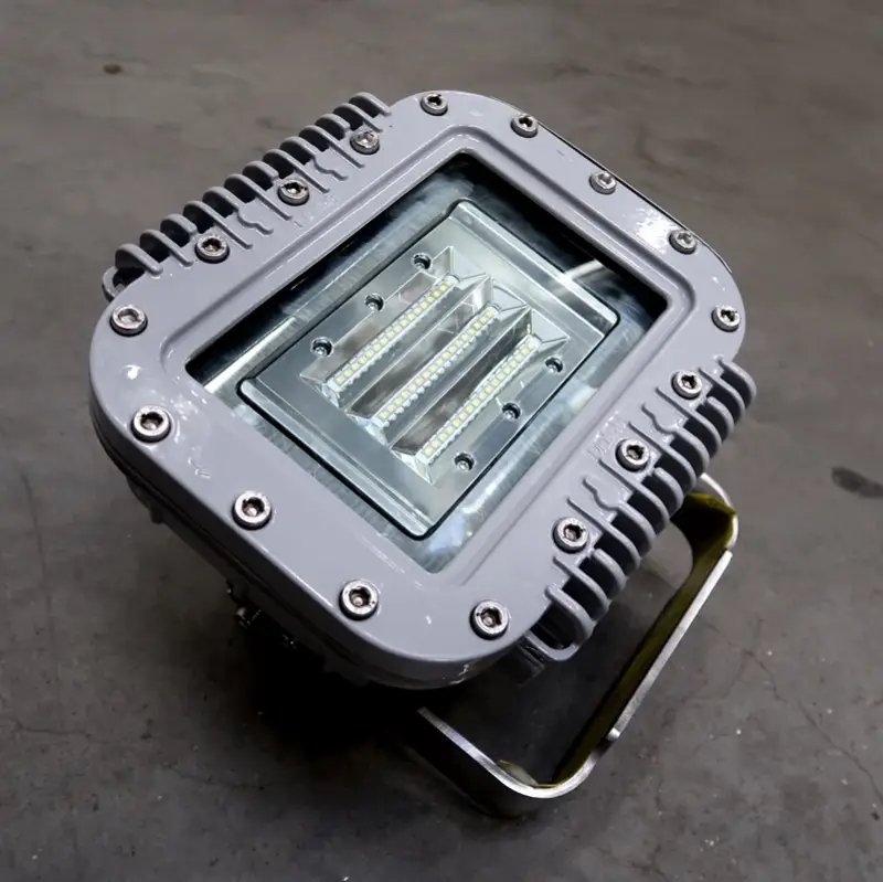 Een grijze industriële LED-schijnwerper met een geribbelde, rechthoekige metalen behuizing en een glazen afdekking, die op een metalen voetstuk op een betonnen vloer staat.