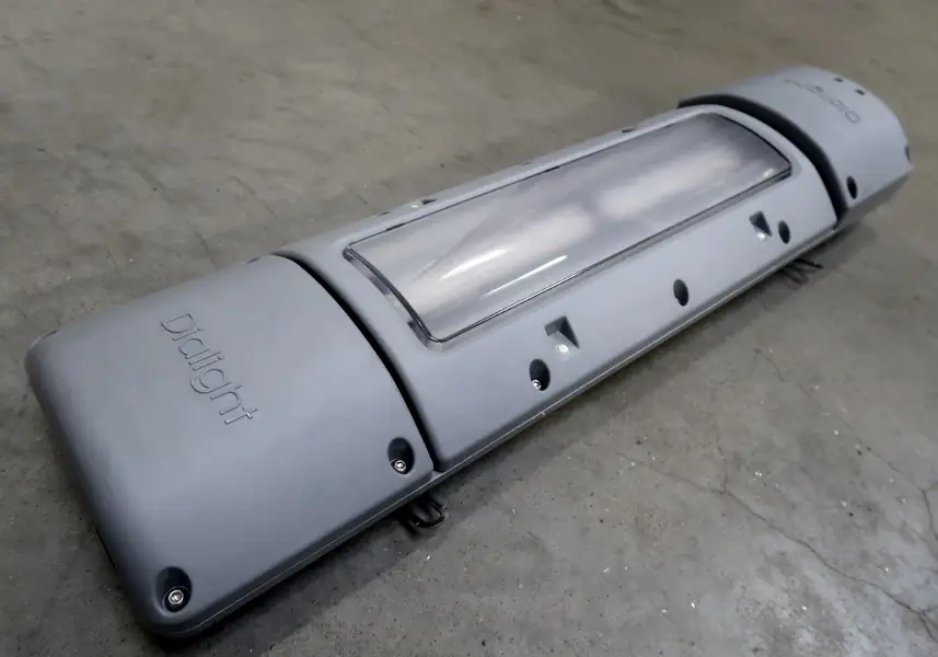 Een grijze Drallight industriële LED-lamp met een doorzichtige kap staat op een betonnen vloer, gezien vanuit een hoek.