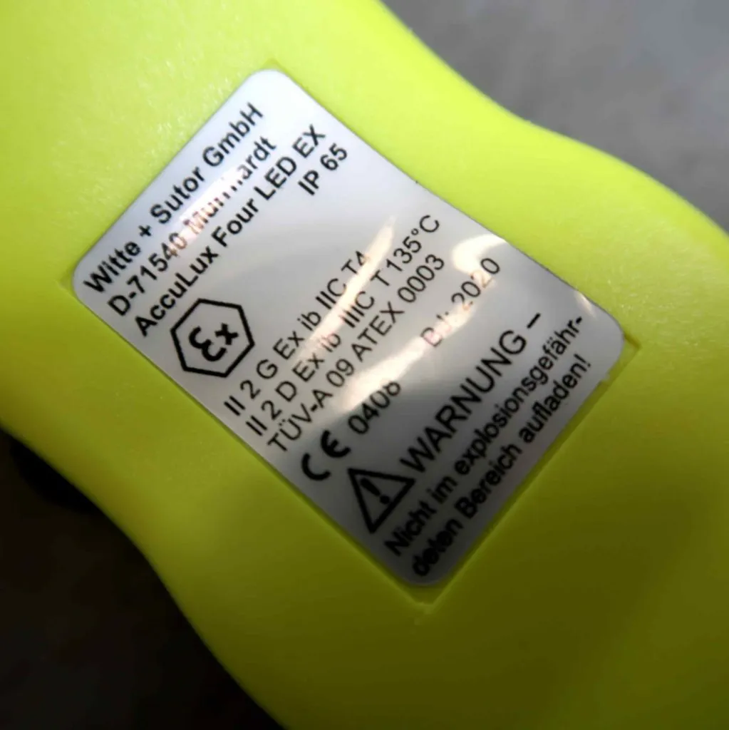 Close-up van een productetiket op een geel apparaat, met technische specificaties, certificeringen en een waarschuwing in het Duits. Op het etiket staan „AccuLux Four LED EX”, „IP65” en „ATEX-conformiteit” vermeld.