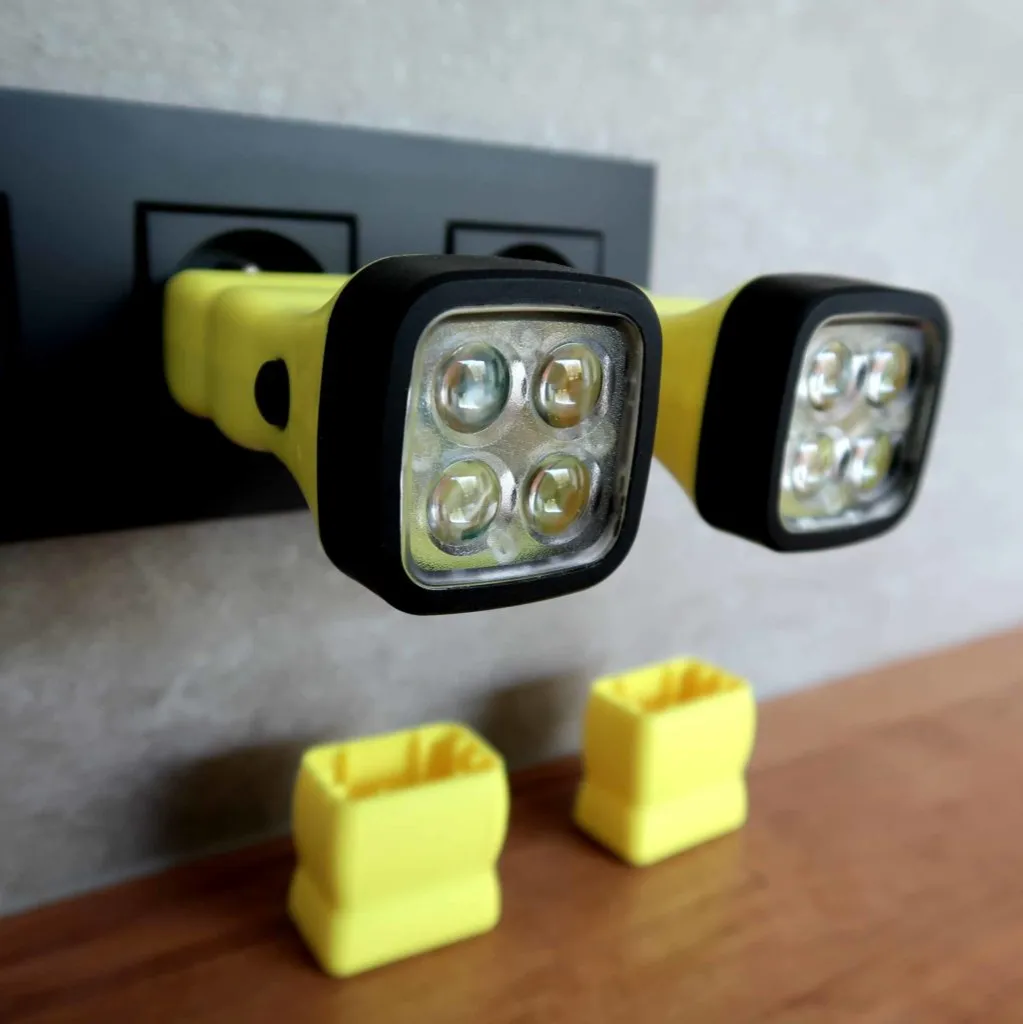 Er zijn twee geel-zwarte LED-zaklampen in een stopcontact gestoken. Onder de zaklampen staan twee lege gele houders op een houten oppervlak. De achtergrond is een lichtgekleurde muur.