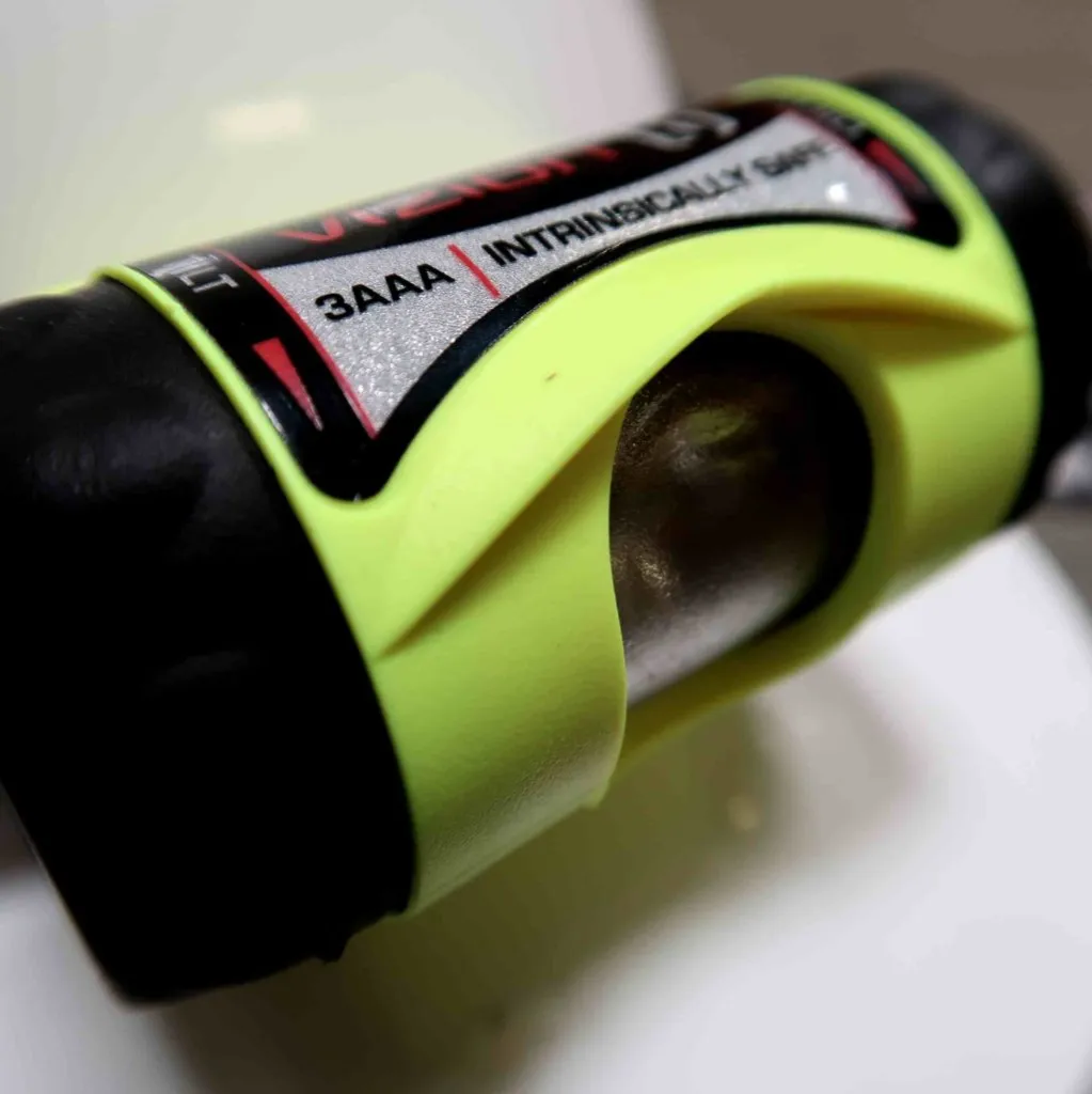 Close-up van een geel-zwarte zaklamp met het opschrift „3AAA INTRINSICALLY SAFE“, waarbij een deel van de lens en de omringende behuizing zichtbaar zijn.