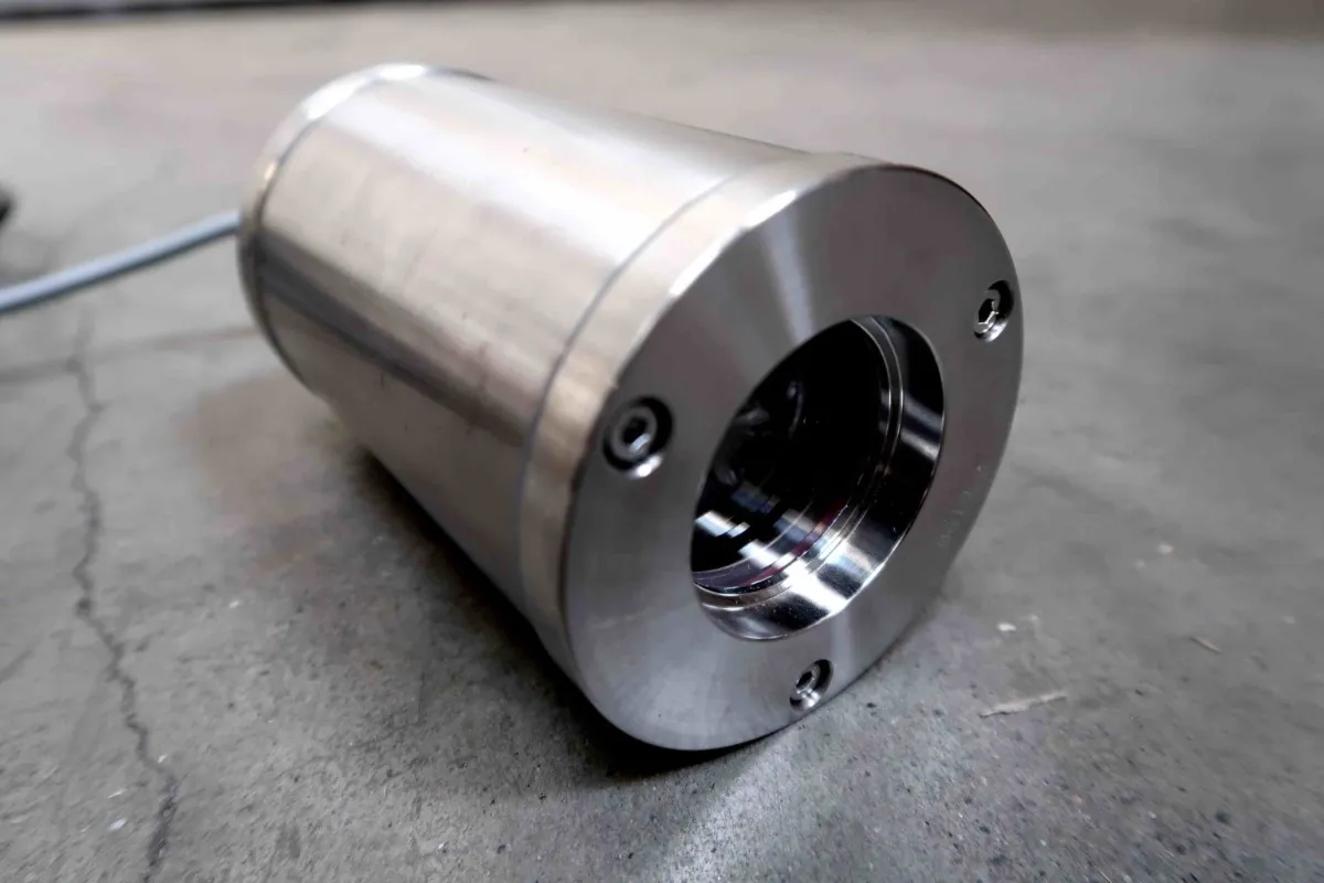Een close-up van een cilindervormige industriële camera of sensor van roestvrij staal die op een grijs betonnen oppervlak ligt, met zichtbare schroeven en een kabel die aan de achterkant uitsteekt.