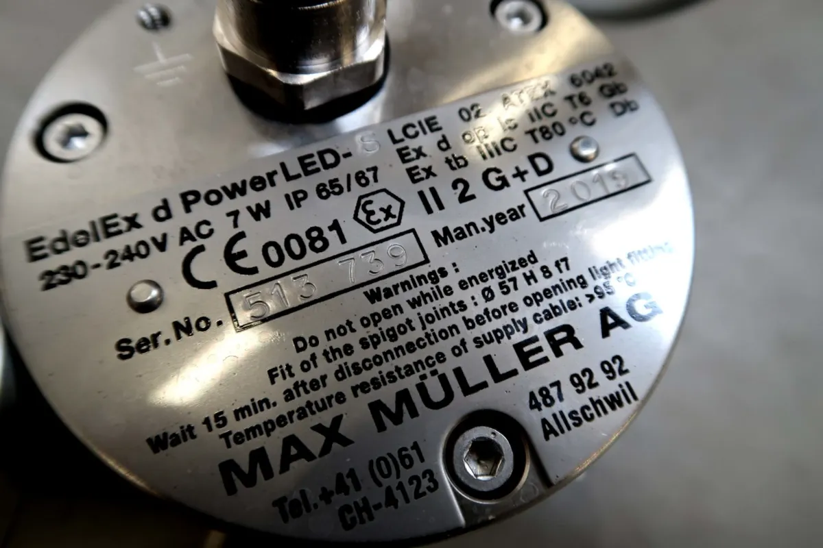 Close-up van een metalen label op een elektrisch apparaat met technische gegevens, modelnummers, veiligheidswaarschuwingen, keurmerken en de gegevens van de fabrikant: MAX MULLER AG, Zwitserland.