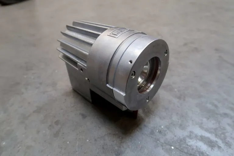 Op een grijs betonnen oppervlak staat een metalen, cilindrisch mechanisch onderdeel met ribben en een ronde opening. Het onderdeel lijkt te zijn vervaardigd uit gefreesd aluminium en heeft een robuust, industrieel ontwerp.